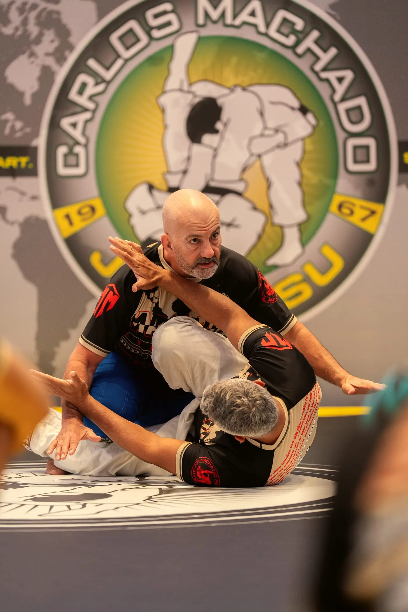 Master Carlos Machado, in a Machado rashguard, controls a partner on the mat, the full Carlos Machado Jiu-Jitsu banner behind