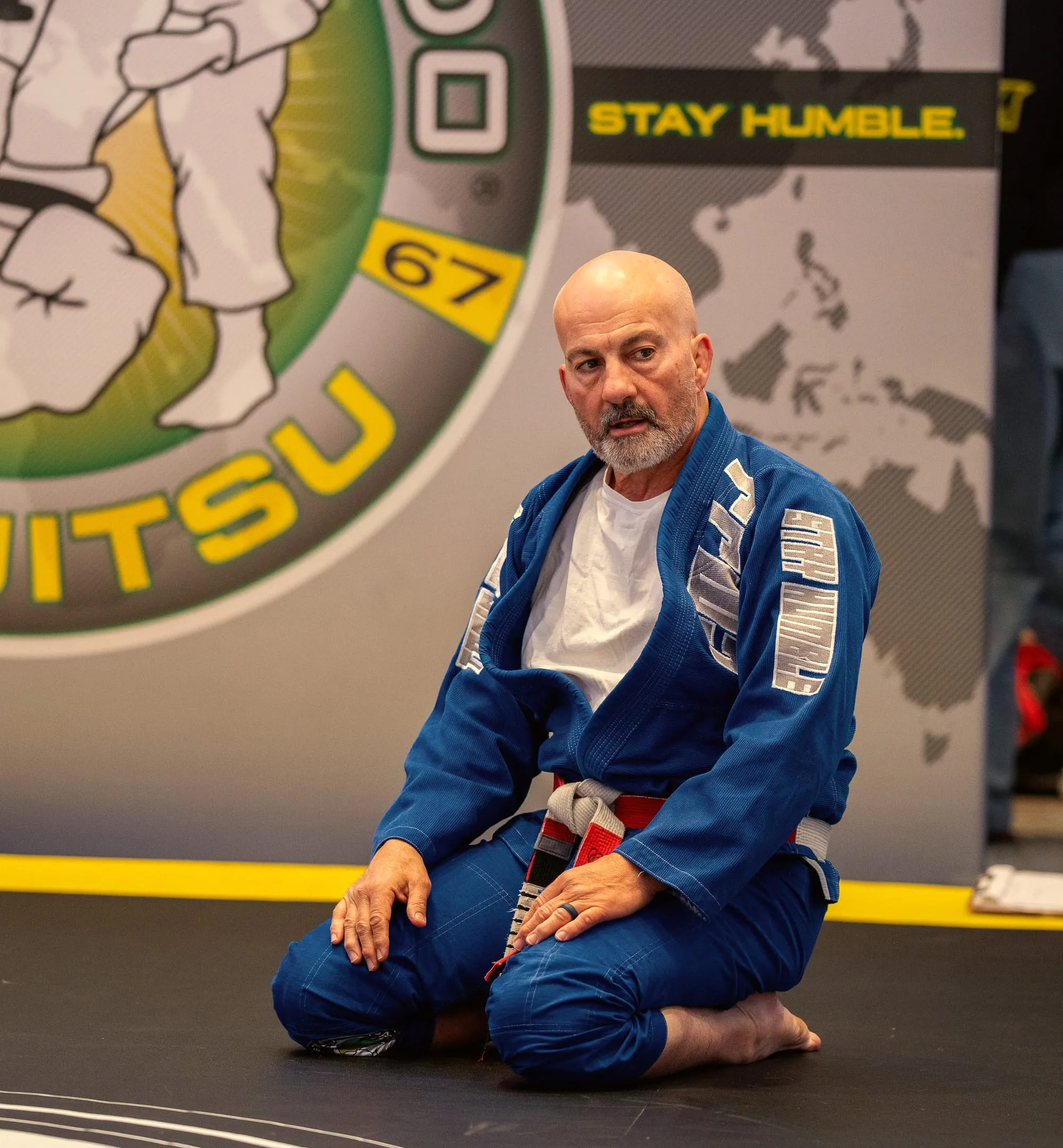 Master Carlos Machado kneels on the mat in a blue gi at the Machado Method Summit, the Carlos Machado Jiu-Jitsu banner behind him