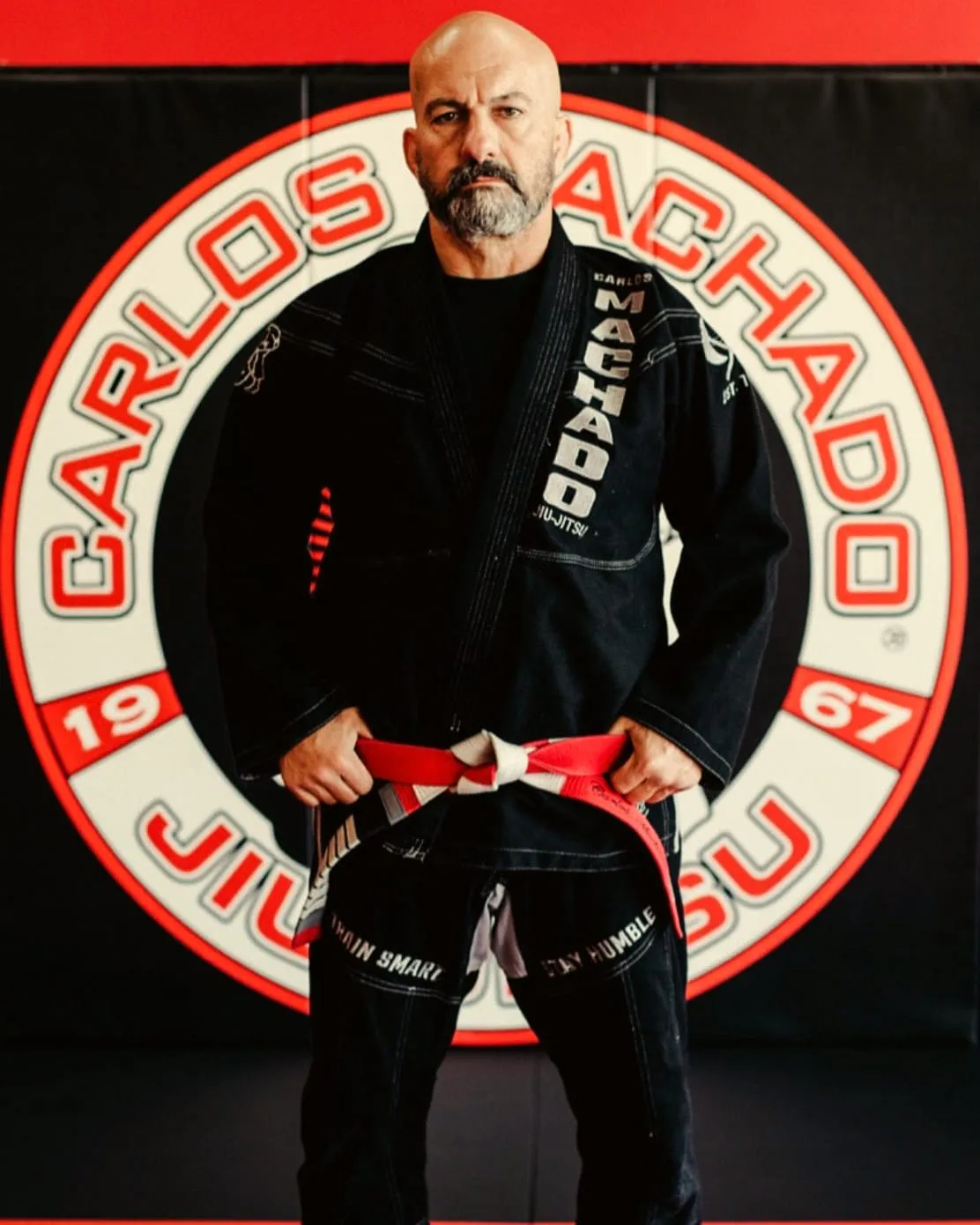 Master Carlos Machado in a black gi and a red and white belt, standing in front of the Carlos Machado Jiu-Jitsu logo