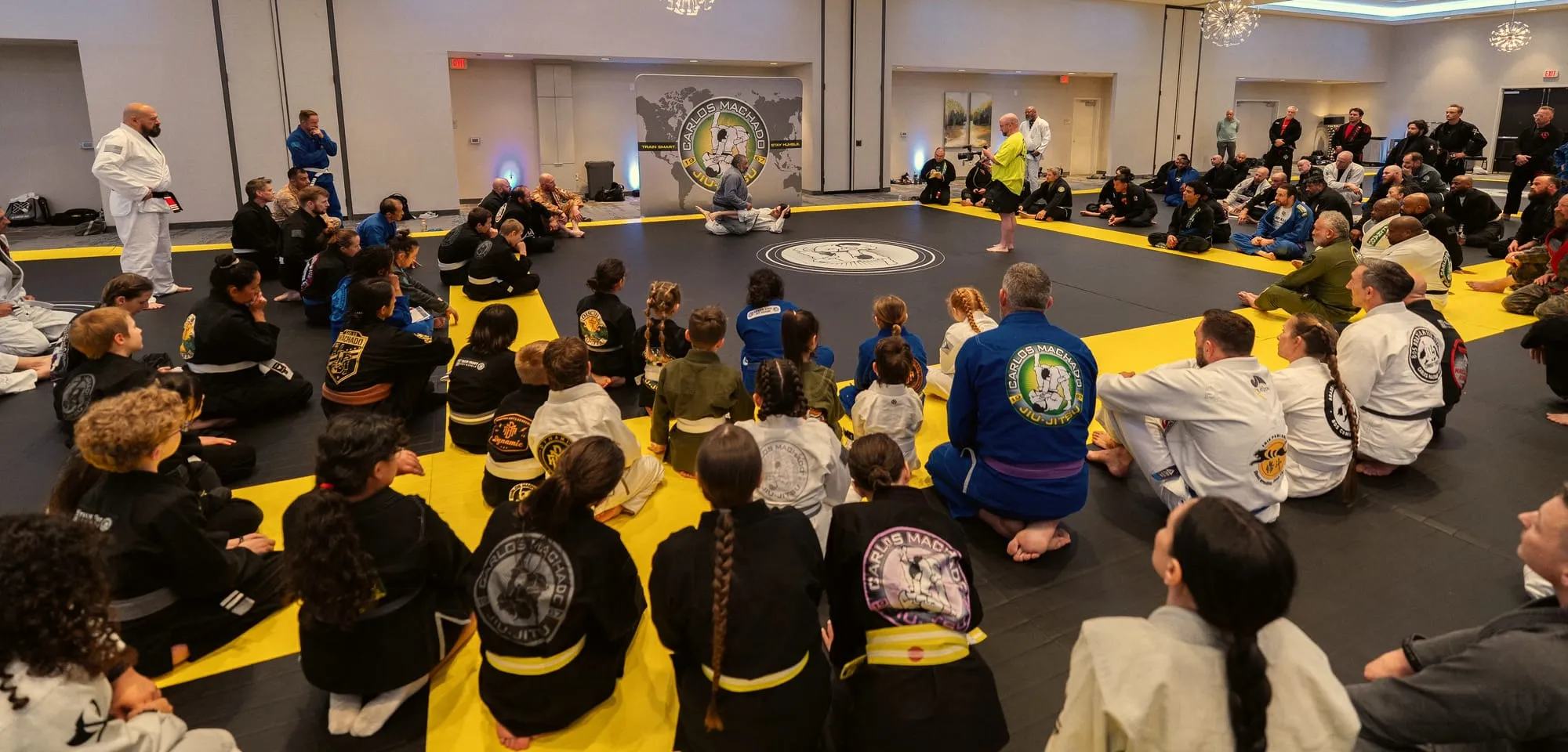 A large seated crowd rings the mat, facing the instructor at the Machado Method Summit