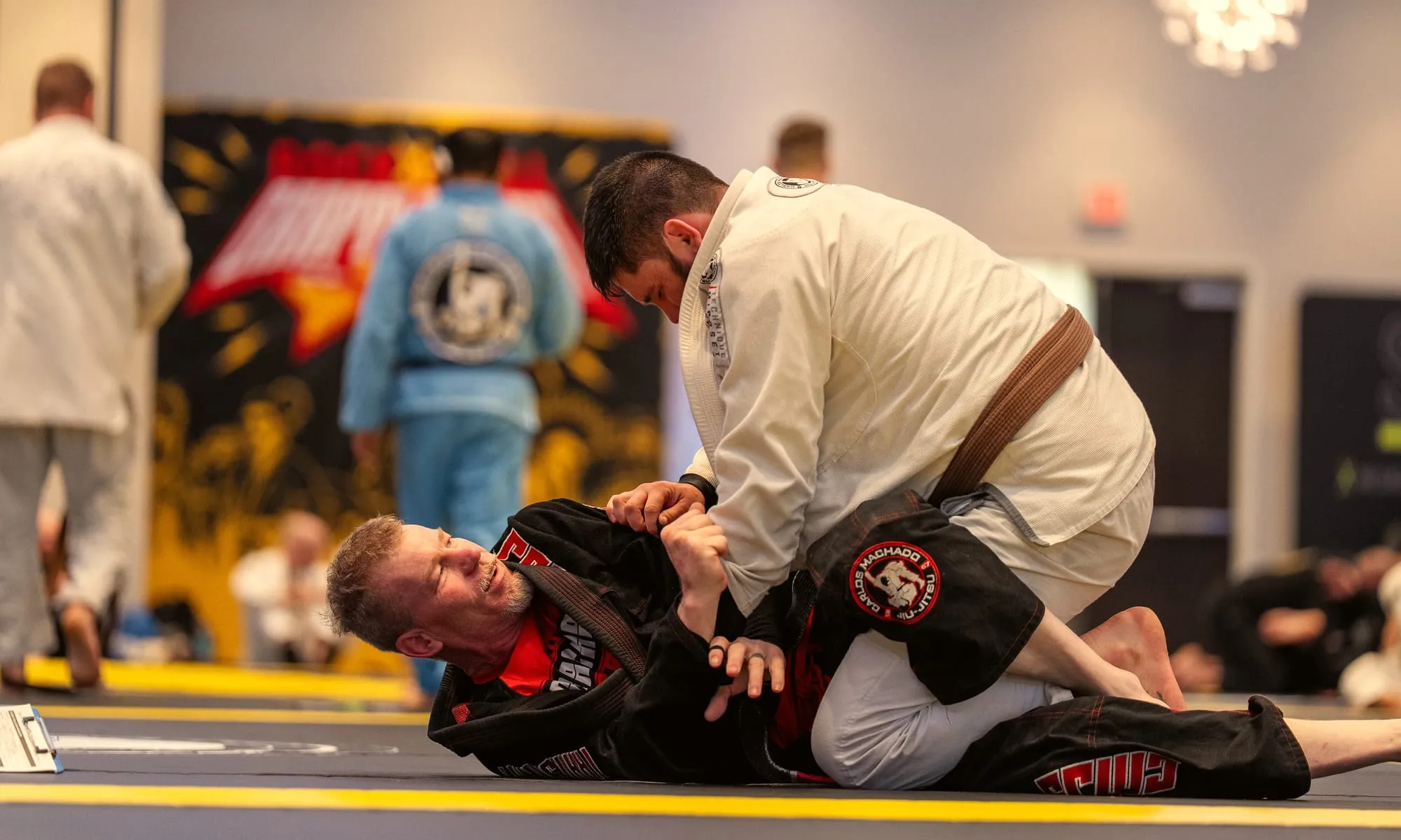 Two students drill a guard pass on the mat, a brown belt in a white gi working over a partner in a black gi