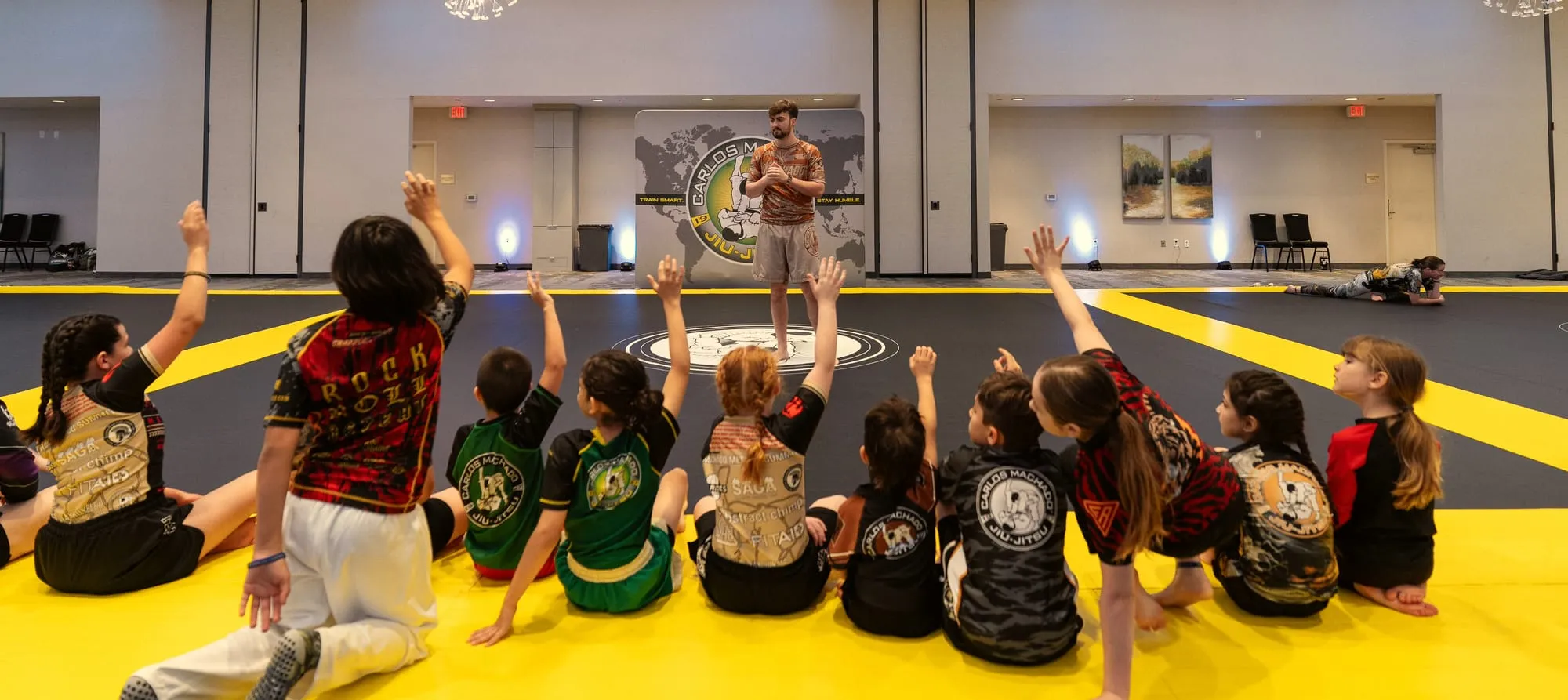 Kids in gis and rashguards sit in a semicircle on the mat with their hands up to answer the coach