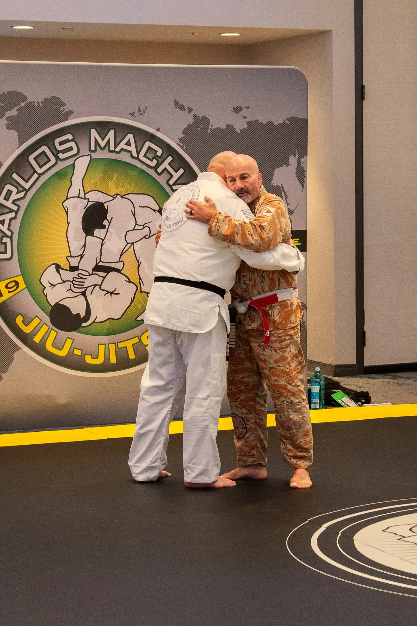Two men embrace on the mat right after a belt promotion, the Carlos Machado Jiu-Jitsu banner behind them