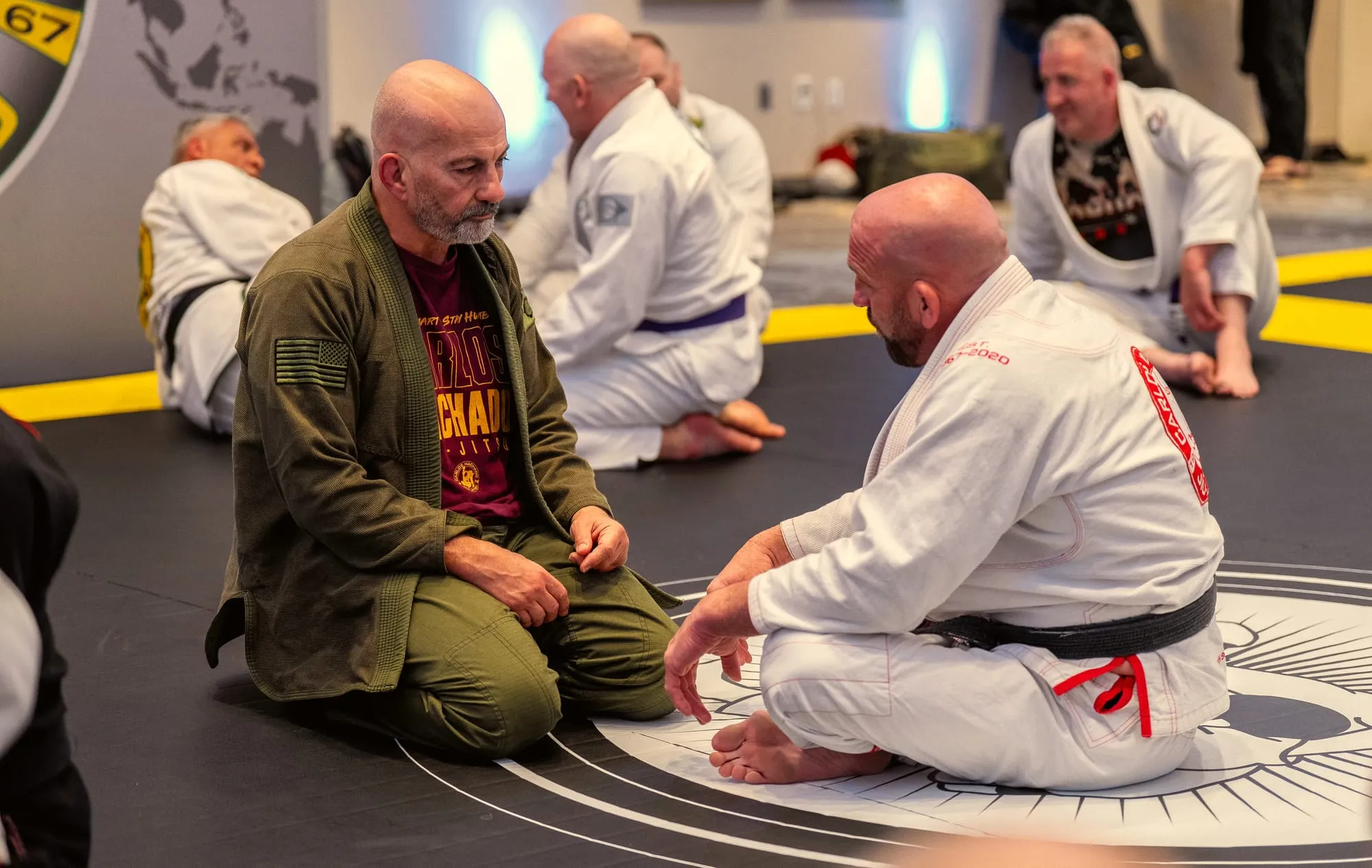 Two black belts kneel face to face on the mat logo during a promotion