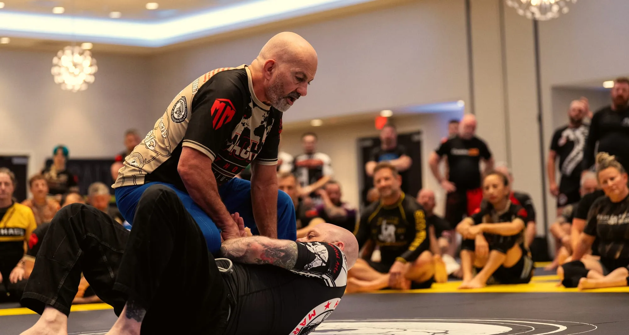A senior instructor demonstrates a hold on the mat while rows of seated adults watch