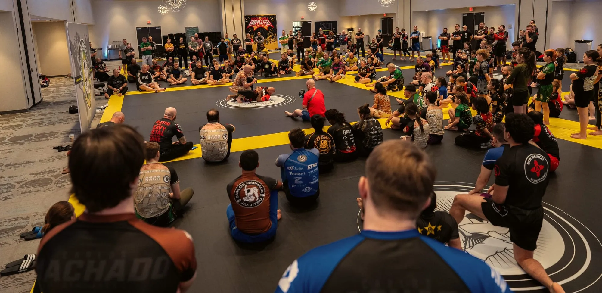 A seated room of students ringing the mat, watching a demonstration at the Machado Method Summit