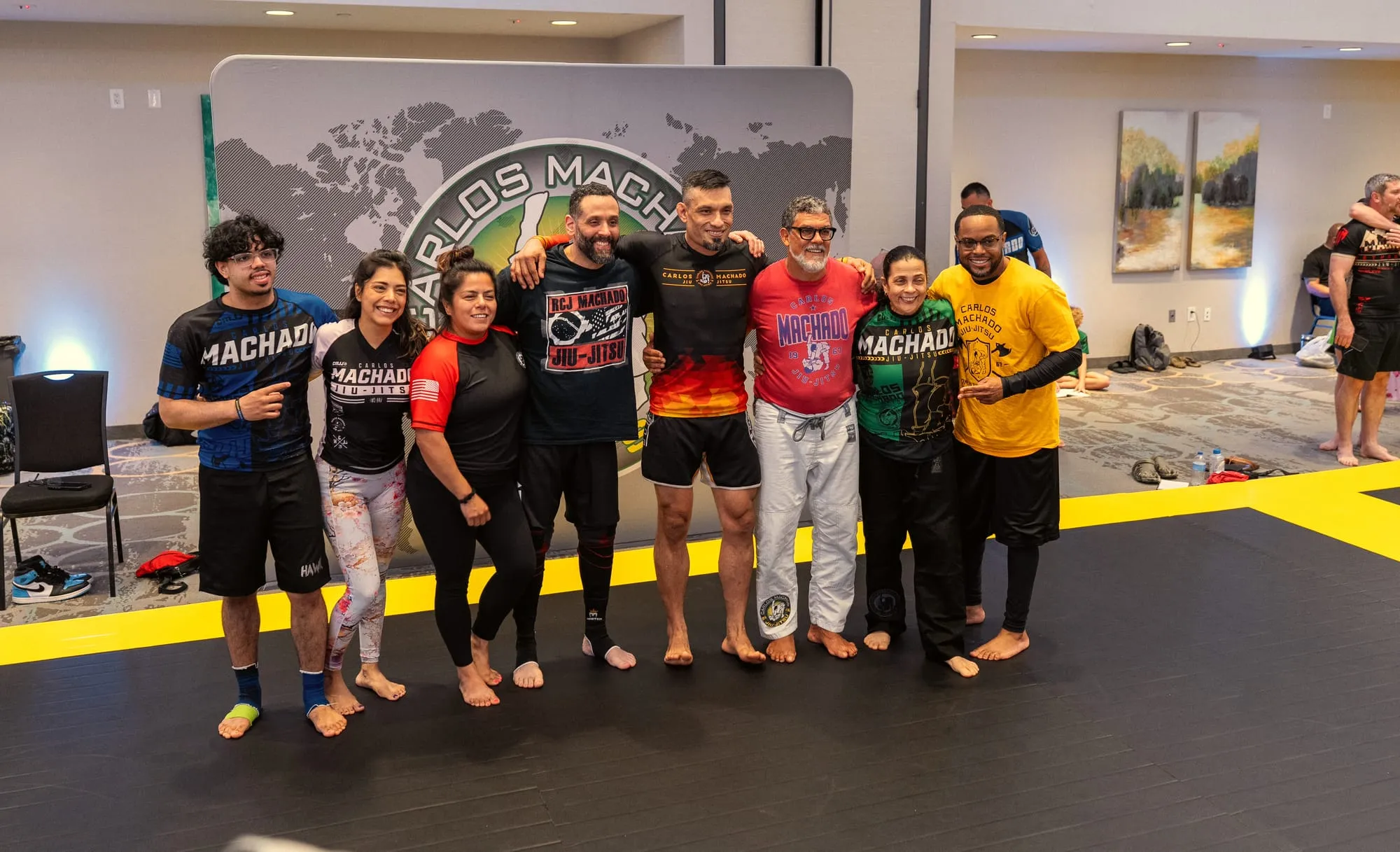 A coaching staff of eight stands together in front of the Carlos Machado Jiu-Jitsu banner