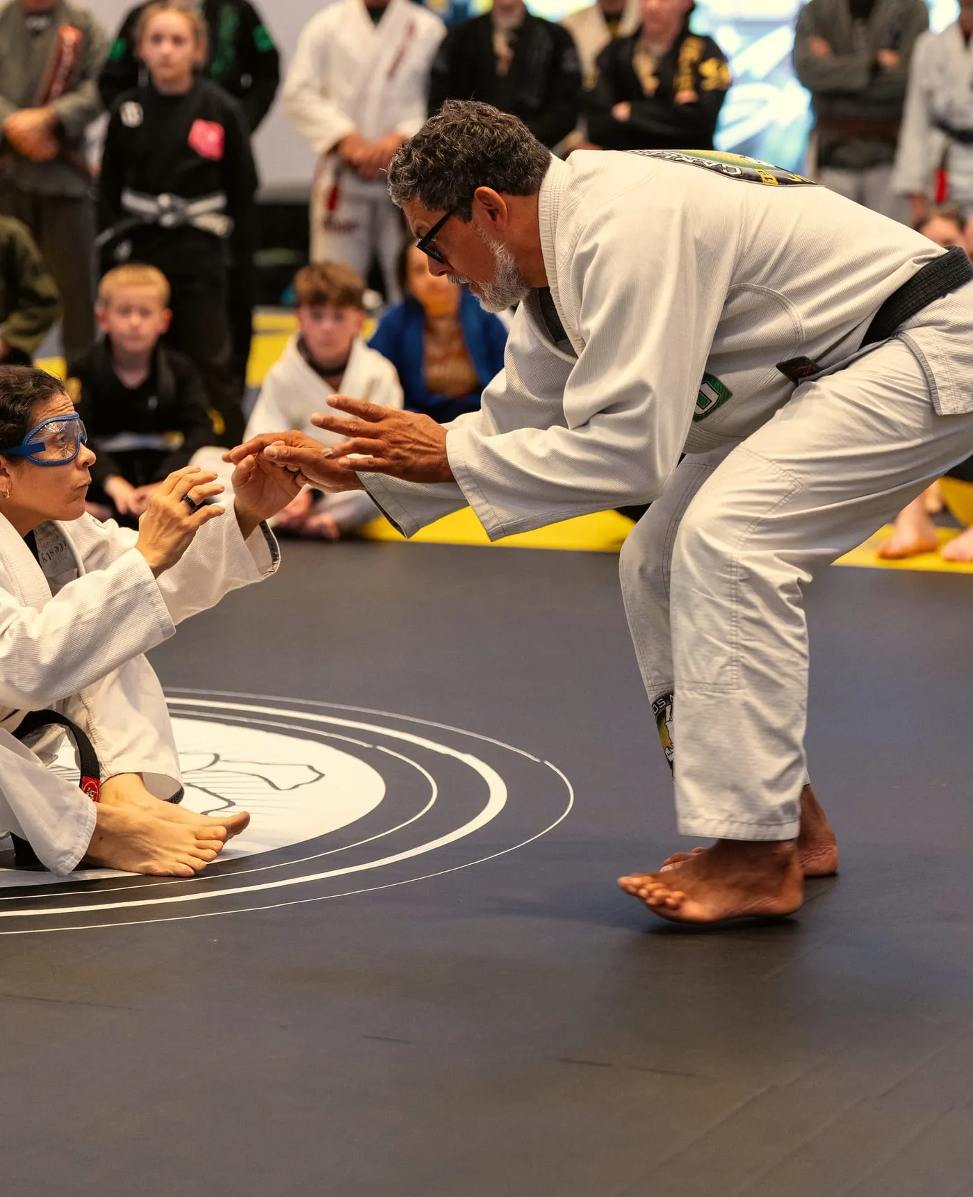 A black belt demonstrates on the mat while kids and adults watch from the edge
