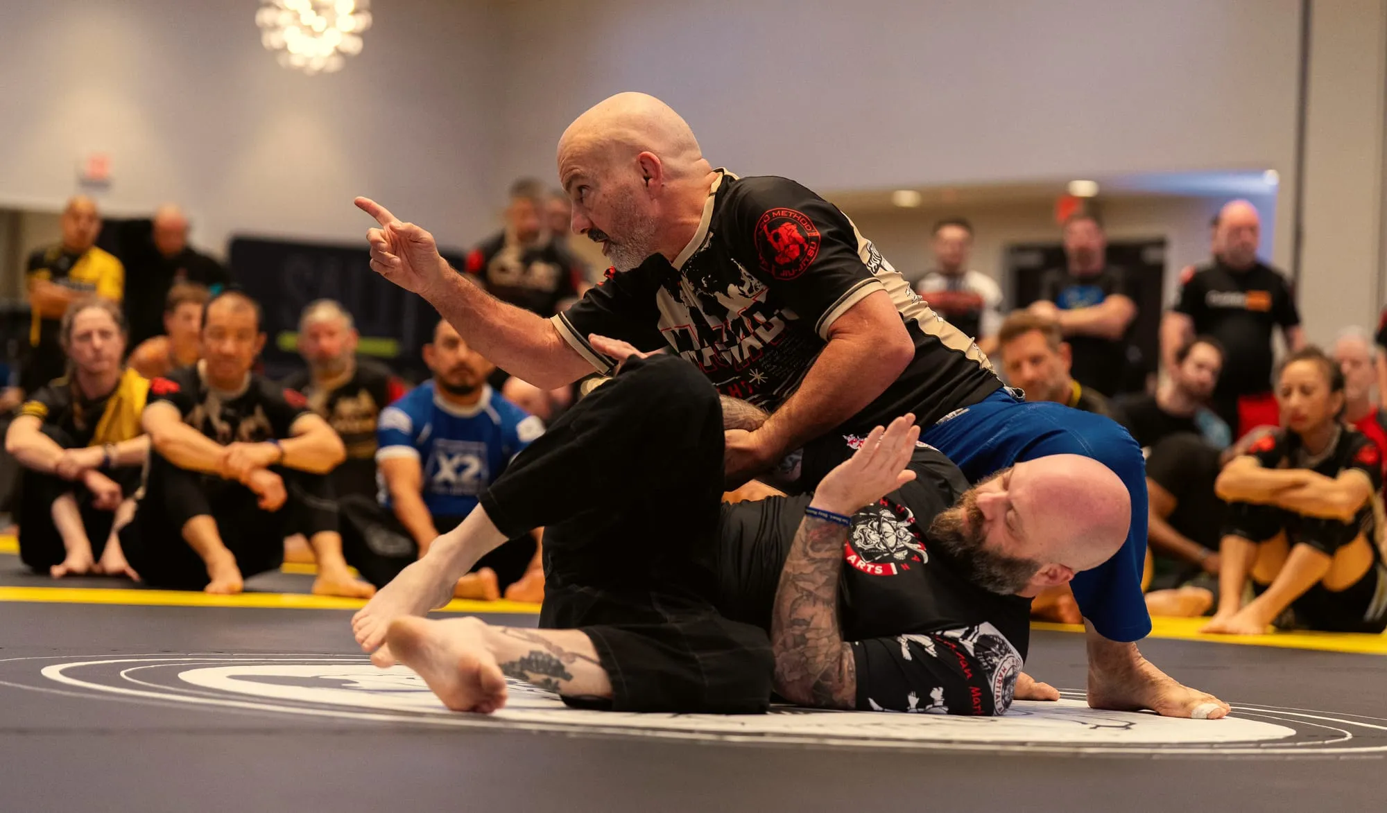 Master Carlos Machado points to make a teaching point while holding a partner down, the room watching from the mat edge